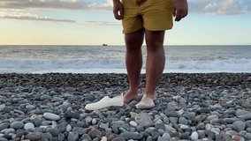 Video 4K. White adult bald man with hairy bare legs in yellow shorts puts on protective transparent shoes on pebble beach against backdrop of sunset, setting sun and sea waves, ocean. Travel, tourism