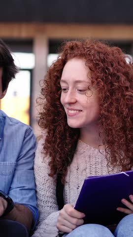 Vertical video of three young international students sitting on the stairs outside the university building. Friends gathered together happily studying with folders, workbooks and notes on campus