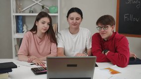 Students communicate using a laptop and the Internet. A guy and two girls look into the laptop camera, smile, wave their hands and read notes from a notepad, sitting at a table in the living room