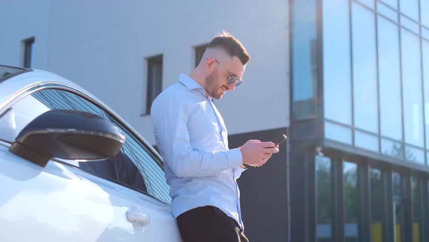 A slender business man in sunglasses is standing looking at the phone next to the expensive car.