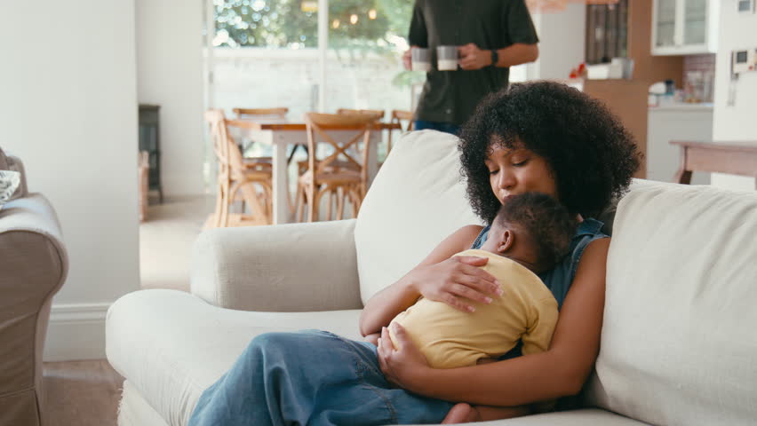 Family with mother cuddling sleeping baby daughter working on laptop sitting on sofa at home as father brings her hot drink - shot in slow motion