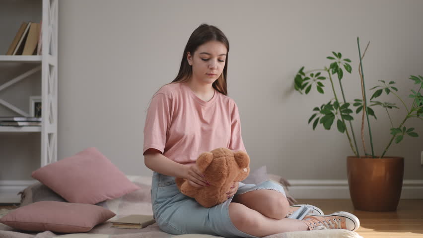 A girl with long hair hugs a teddy bear and looks thoughtfully into the distance, sitting on a plaid on a wooden floor in the living room