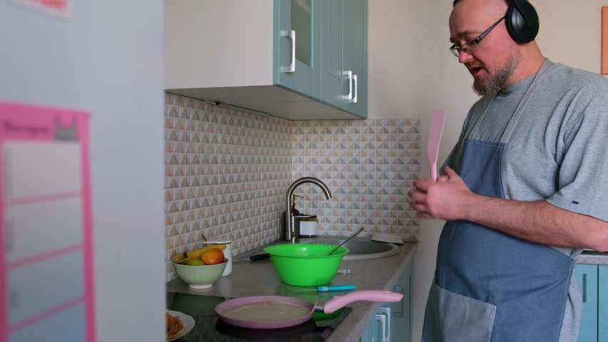 Adult caucasian man cooking breakfast in the home kitchen and listening music in headphones. Bald bearded guy wearing apron having fun while baking pancakes on the stove. Royalty-Free Stock Footage #3473681073