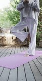 Vertical video of focused biracial woman practicing yoga on sunny terrace, slow motion. Lifestyle, wellbeing in nature, yoga, fitness, healthy lifestyle and domestic life, unaltered.