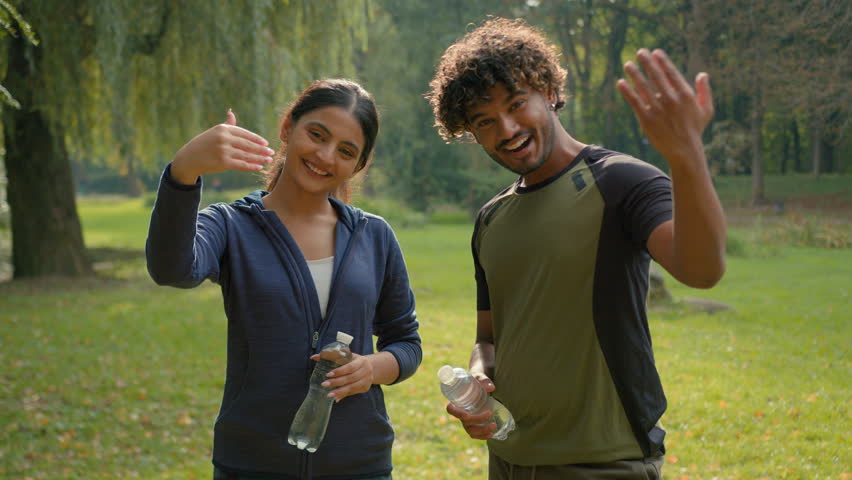 Two sport people smiling talking holding water bottles smile invitation join to exercise male Indian female Arabian friends couple man woman in park invite welcome workout fitness fit in city motivate