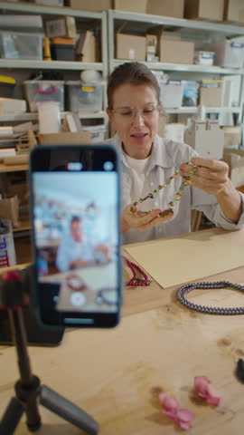 Vertical rack focus shot of woman showing handmade jewelry and telling about it on camera in workshop while filming video advertising or DIY vlog with smartphone on tripod Royalty-Free Stock Footage #3473829677