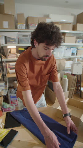 Vertical shot of young man packing garment into cardboard parcel box while preparing order for shipping to customer in online store warehouse