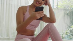 Tilt up shot of cheerful Asian woman in earphones watching funny videos on mobile phone while resting after home workout