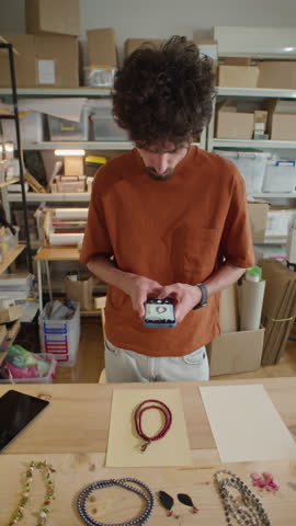 Vertical shot of young man standing by desk in online store warehouse and taking photos of handmade necklaces on beige background with smartphone for sale