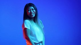 Positive smiling young, beautiful Asian woman dressed casual outfit posing looking at camera against blue gradient background in neon light. Concept of human emotions, self-expression, fun and joy.