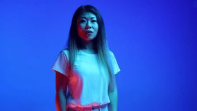 Surprised young woman with open mouth looking at camera and holds hands raised against blue gradient background in neon light. Concept of human emotions, facial expression, self-expression.