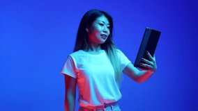 Smiling Asian woman waving hello while holding video meeting in digital tablet against blue gradient background in neon light. Concept of business, startup, sales, working and studying in distance.