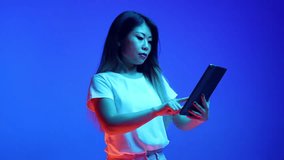 Young, attractive Asian woman using digital tablet against blue gradient background in neon light. Working in a distance. Concept of business, study, work, startup, sales, shopping, delivery services.