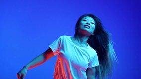 Overjoyed, relaxed Asian woman dancing raising hands against blue gradient studio background in neon light. Concept of human emotions, self-expression, music and dance, fun and joy.
