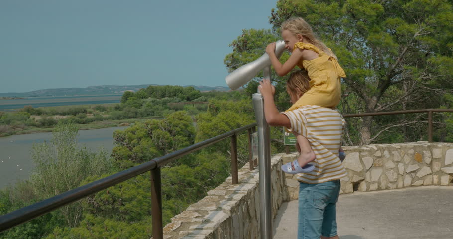 A boy in a striped shirt lifts a girl in a yellow dress, onto his shoulders so that she can look through a stationary open telescope at a pond with distant hills
