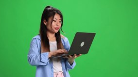 Young Asian woman looking to digital laptop with shocked facial expression against vibrant green studio background. Concept of business, study and work, startup, sales season, shopping.