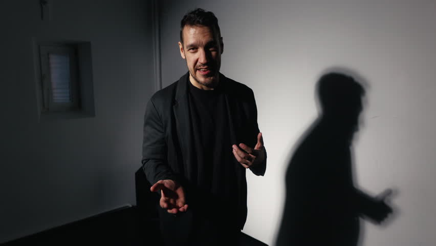 A suited man with a shadow on the wall gestures with his thumb in a dark room