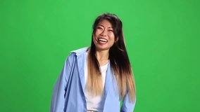 Young beautiful Asian woman, student in casual outfit laughing looking at camera against vibrant green studio background. Concept of human emotions, self-expression, fashion, style.