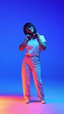 Joyful young Asian woman in casual attire dancing raising hands against blue gradient background in neon light. Concept of human emotions, self-expression, youth and beauty, dance and music.