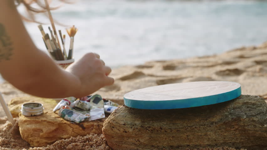 Female artist prepares color palette by seaside at sunset for painting. Creative setup on sandy shore near waves. Hands mixing pigments, arranging brushes preparing art project on beach. Slow motion.