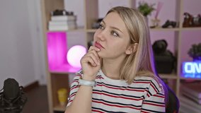 Young, blonde streamer woman thinks doubtfully, a pensive smile playing on her face as she ponders the gaming question, hand on chin, in a dark room