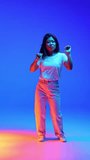 Joyful young Asian woman in casual outfit dancing raising hands against blue gradient background in neon light. Concept of human emotions, self-expression, dance and music, fun and joy. Ad