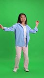 Young attractive Asian woman, student energetically dancing raising hands in casual outfit against vibrant green studio background. Concept of human emotions, self-expression, fun and joy, happiness.