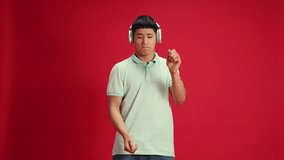 Overjoyed young Asian man in casual attire dancing of rejoice while listening favorite music against vibrant red studio background. Concept of human emotions, fashion and beauty, self-expression, work