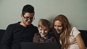 Happy parents and their teenager watching videos and photos in the family album on the computer. Modern young family sitting on the couch and using the laptop at weekends. Concept of memories.