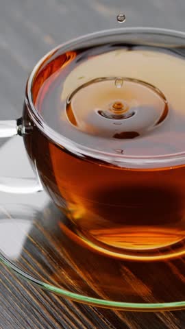 VERTICAL VIDEO: Drops of fragrant black tea fall into transparent cup with tea. Slow motion shot of tea splash, Pouring tea into cup
