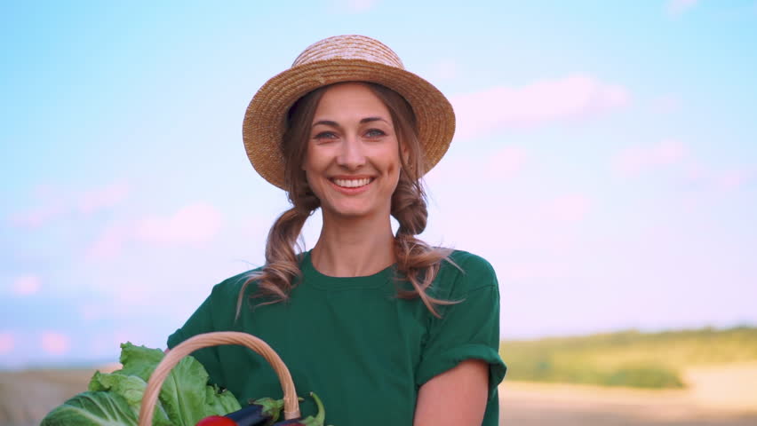 Woman farmer in straw hat holding basket full of vegetables standing on farmland. Portrait of smiling female agronomist specialist farming agribusiness looking at camera.