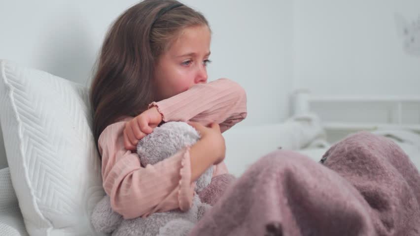 Upset sick girl coughing while sitting with soft toy on bed at home. Sad girl child suffering from cold and flu and using digital tablet in apartment.