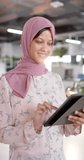 Vertical video of happy biracial casual businesswoman in hijab using tablet in office, slow motion. Casual office, business, communication and work, unaltered.