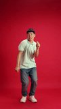 Funny, cheerful, young Asian man, student in casual attire dancing with joy in motion against red studio background. Concept of emotions, fashion and beauty, self-expression, work and hobby.