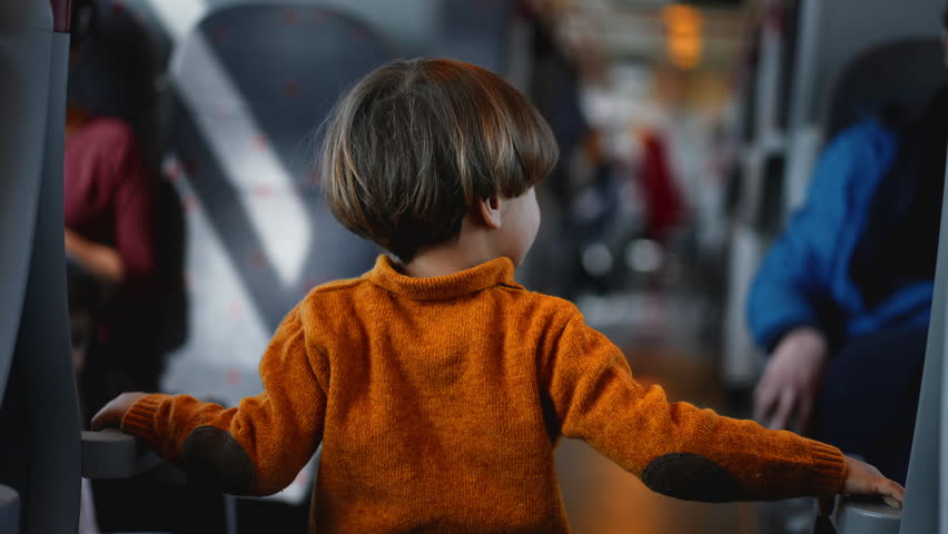 Brightly dressed lad in a yellow sweater strolls the train aisle, gripping the seat handles for support. The junior traveler spends his commute time meandering aboard the rail-car Royalty-Free Stock Footage #3474056003