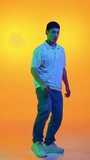 Funny, cheerful, young Asian man dancing with joy in motion in vibrant neon light against gradient orange background. Concept of human emotions, fashion and beauty, work and hobby, fun and joy.