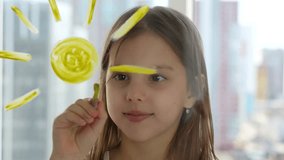 Cute girl painting sun on window with paintbrush, close-up. Little girl painting sun with paintbrush and yellow paint on glass surface, 4k
