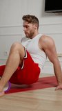 Vertical video Confident happy athlete man blond with stubble in a white T-shirt and red shorts doing exercises and massaging his thigh using a special roller massager in a modern apartment in the