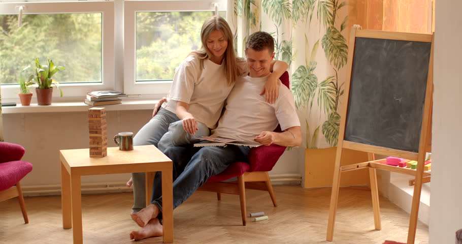 Man and woman reading book. Warm and inviting ambiance of nursery, young parents sit side by side, immersed in nostalgic journey of flipping through a photo album. Use printed publications on paper.  Royalty-Free Stock Footage #3474111907