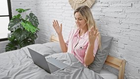 A young blonde woman in pink pajamas engages in a friendly video call from her cozy bedroom with a laptop and a decorative wall background.