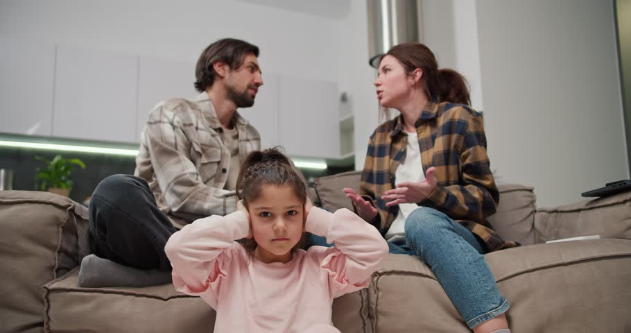 Little brunette girl in pink clothes covers her ears and is sad while her parents are quarreling while sitting on the sofa in a modern apartment. A man with stubble in a beige T-shirt is quarreling