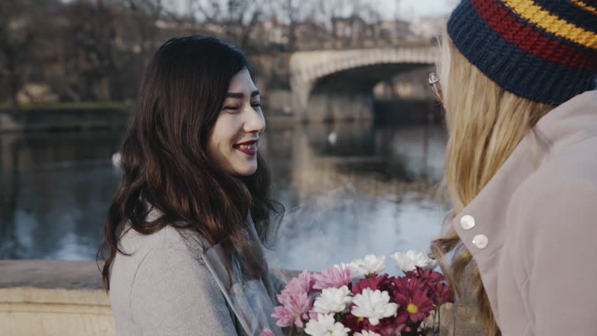 Close-up portrait of happy lesbians on date. Blonde girl giving asian girlfriend bunch of flowers. Young homosexual couple spending time together on holiday of valentine day. Romantic, LGBTQ concept.