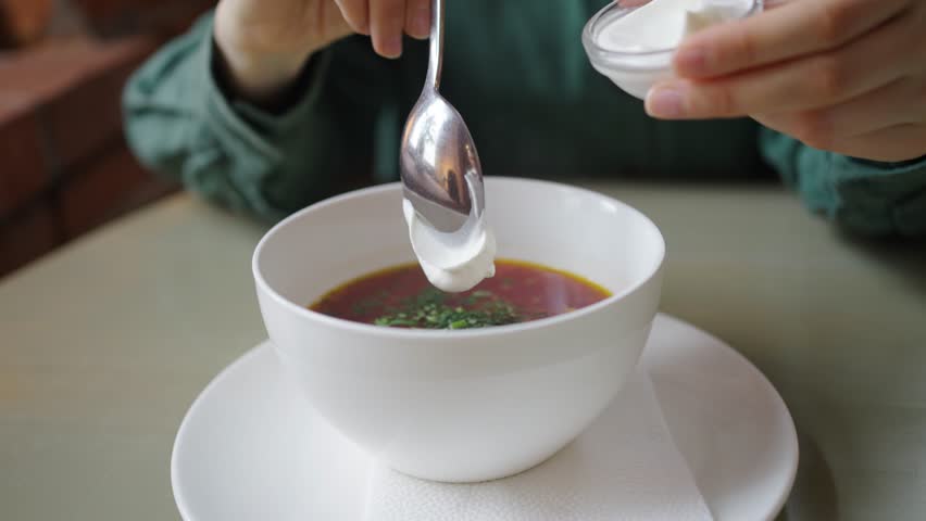 Woman gently adds a dollop of sour cream to a bowl of vibrant borscht, garnished with fresh herbs, in a well-lit setting perfect for culinary themes. Healthy Organic Borscht Soup Being Seasoned with
