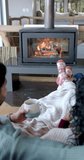 Vertical video of biracial couple sitting on sofa in front of fireplace at home, slow motion. Lifestyle, relaxation and domestic life, unaltered.