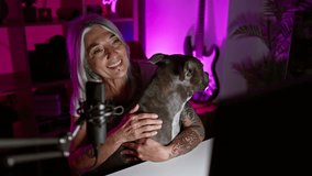 Smiling, confident grey-haired middle age woman streamer with pet dog sparks joy while speaking, playing games in a cozy gaming room, captivating digital world with her mature charisma