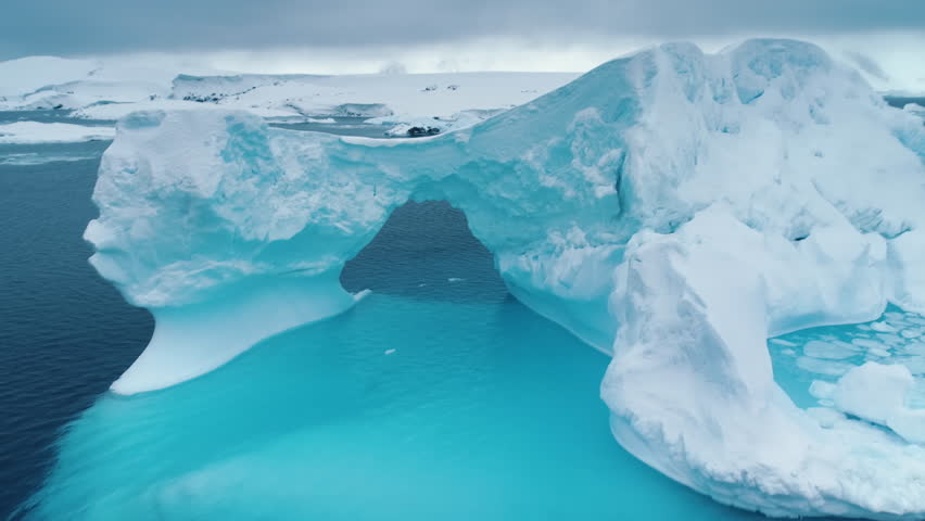 Large iceberg floats in Antarctica ocean, huge towering frozen arch cave with blue ice melting underwater. Climate change at polar glaciers winter landscape. Aerial drone footage zoom out panorama