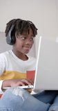 Vertical video of happy african american boy sitting on bed using laptop at home, slow motion. Childhood, communication and domestic life, unaltered.