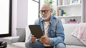 A mature man with glasses and a beard reads on a tablet in a well-lit home living room, conveying a relaxed atmosphere. - Powered by Shutterstock - Get 15% off with code: PIKWIZARD15