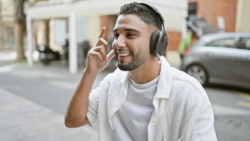 Handsome young man with beard enjoys music on headphones in urban city street setting.