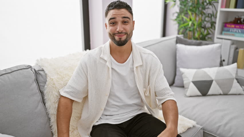 Cheerful young arab man grinning wide, open arms beckoning for a hug on camera in his living room